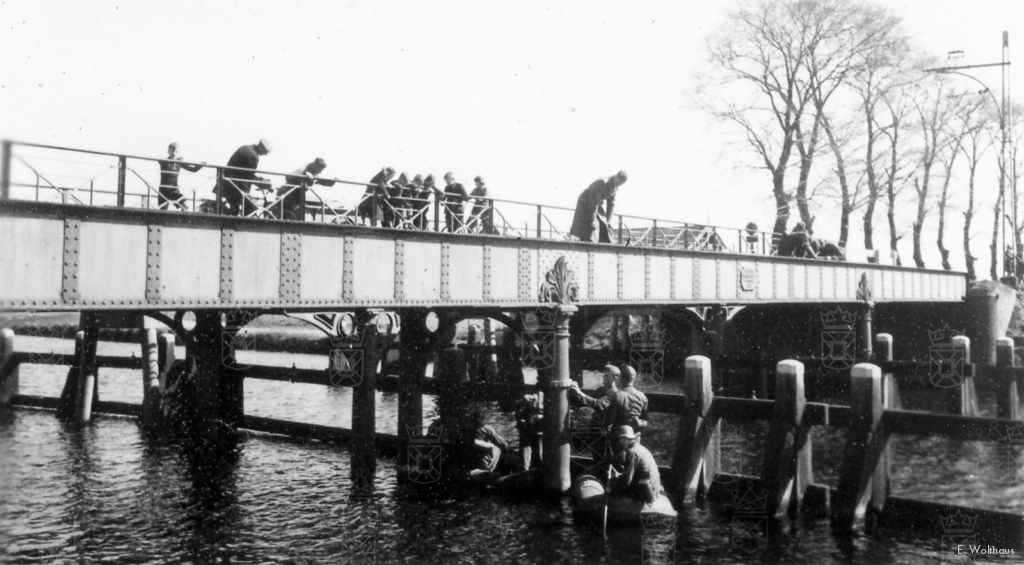 Duitse militairen bevestigen vanuit een rubberbootje een springlading aan een pijler van de trambrug. Dit alles onder het toeziend oog van enkele omstanders.